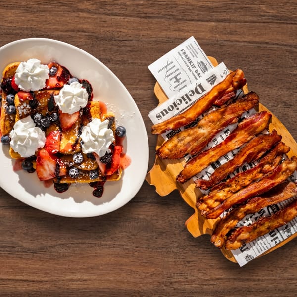 French toast with berries and whipped cream next to a board of crispy bacon on a wooden table.