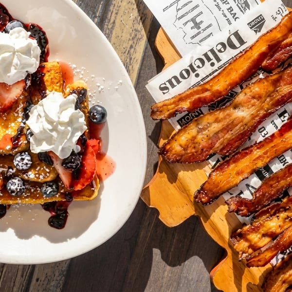 French toast with berries and whipped cream, alongside crispy bacon strips on a wooden board.