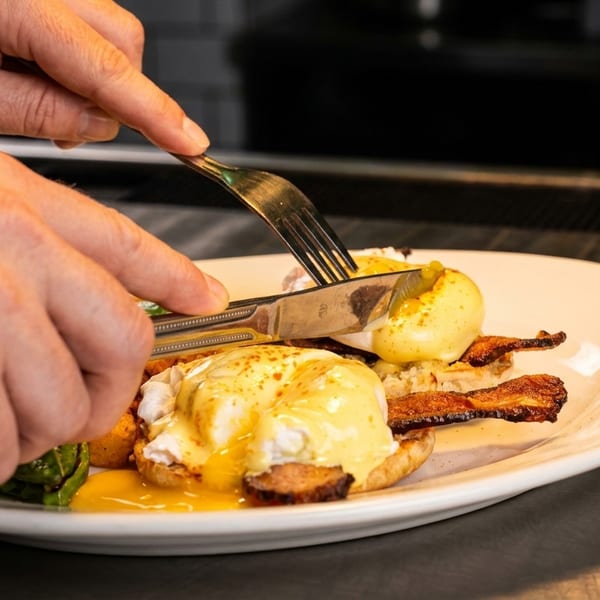 Hands cutting into a plate of eggs Benedict with bacon and roasted potatoes.