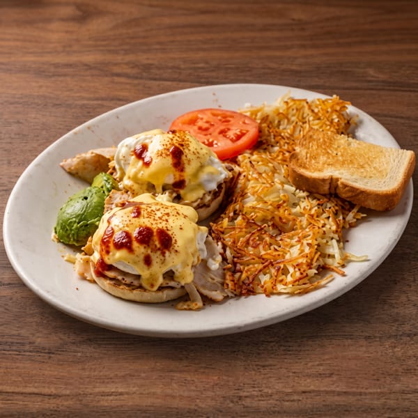 Plate of Eggs Benedict with hash browns, toast, tomato, and mashed avocado.